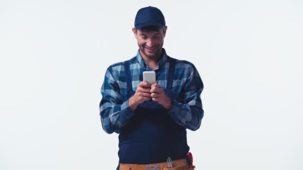 快乐的水管工 身穿制服 头戴帽子 用智能手机发短信 与白色隔离 — 图库视频影像