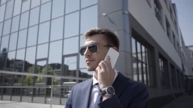 Güneş gözlüklü gülümseyen iş adamı dışarıda akıllı telefondan konuşuyor. 