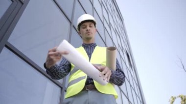 İnşaatçının sabit şapkalı düşük açılı görüntüsü binanın yakınındaki planlara bakıyor 