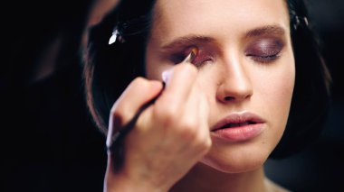 makeup artist applying eye shadow with cosmetic brush on model with closed eyes clipart