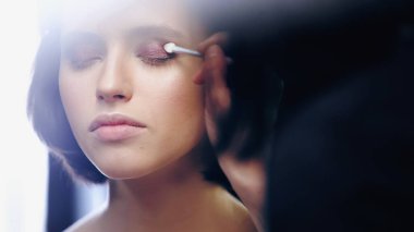 makeup artist applying eye shadow with cosmetic brush on woman with closed eyes clipart