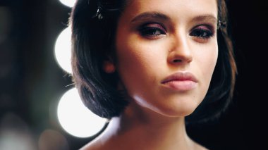 brunette young woman with eye makeup looking at camera with blurred backlit on background  clipart