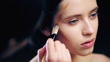 Bulanık makyaj sanatçısı kozmetik fırçayla genç modelin yüz hatlarını çiziyor.