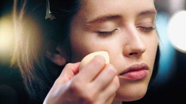 makeup artist applying makeup foundation with cosmetic sponge on face of model with closed eyes clipart