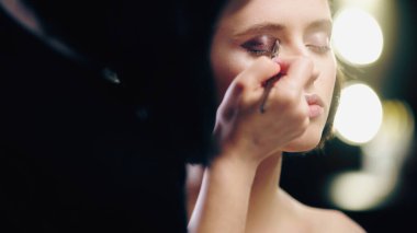 blurred makeup artist applying eye shadow with cosmetic brush on eyelids of model with closed eyes clipart