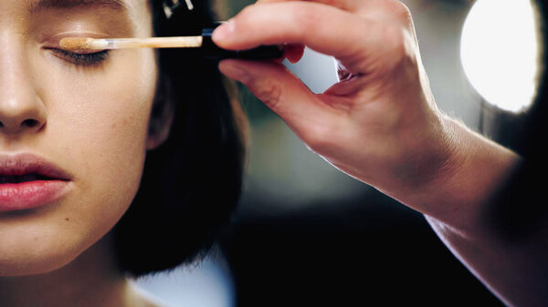 cropped view of makeup artist applying concealer on eyelid of model 