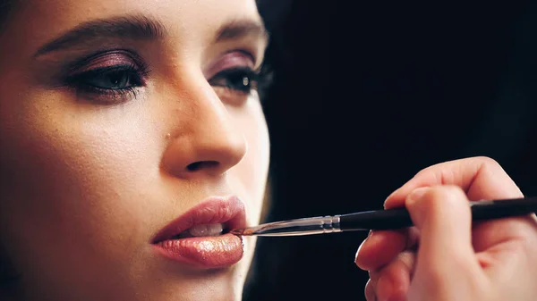 makeup artist applying lip gloss on lips of model isolated on black