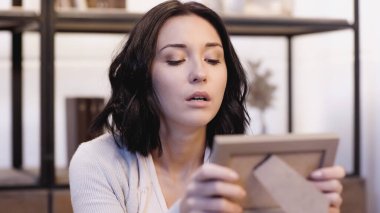 offended brunette woman looking at blurred photo frame in hands at home clipart
