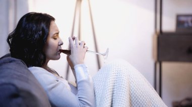 Battaniyeye sarılmış üzgün bir kadın kanepede oturuyor ve evde camdan kırmızı şarap içiyor.