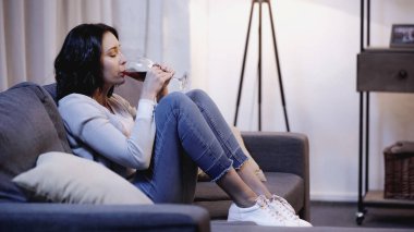 depresif yalnız kadın gündelik giysiler içinde kanepede oturuyor ve evde camdan kırmızı şarap içiyor
