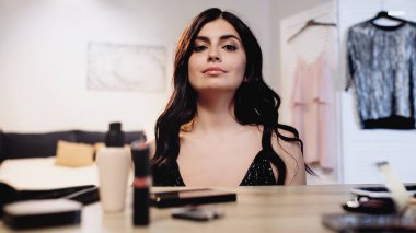 Yatak odasında dekoratif kozmetik ürünleri olan, masasının yanında parlak saçlı mutlu esmer bir kadın.