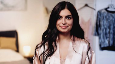 Pembe ipekli, uzun saçlı ve yatak odasında kameraya bakan gülümseyen genç kadın.