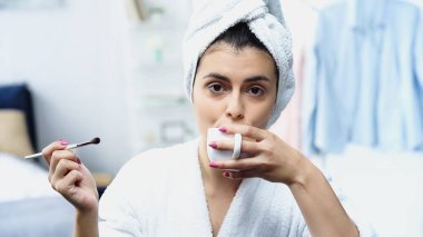 Kafası havluya sarılı genç bir kadın kozmetik fırça tutuyor ve yatak odasında kahve içiyor.