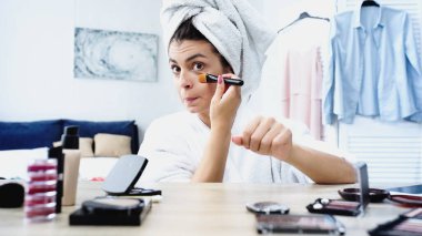 Kafası havluya sarılı genç bir kadın yüzünü buruşturuyor ve yatak odasında makyaj fırçasıyla göz altına alıyor.