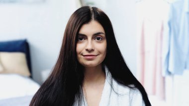 Uzun düz saçlı, bornozlu ve yatak odasındaki kameraya bakan gülümseyen genç kadının portresi.