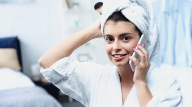 Kafası havluya sarılı genç bir kadın kozmetik fırça tutuyor ve yatak odasında cep telefonuyla konuşuyor.