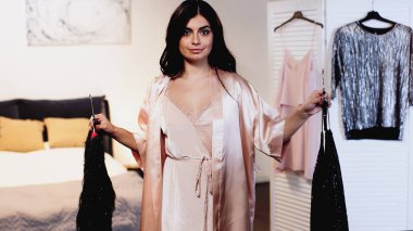 Pembe ipek peignoir giymiş genç bir kadın. Elinde siyah elbiseli askılar var.