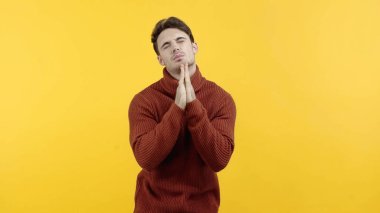 Man in sweater standing with closed eyes and praying hands isolated on yellow  clipart