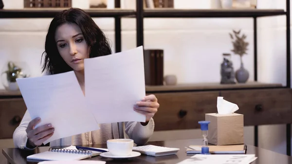 Ciddi bir kadın, elinde fincan kahveyle masada oturuyor ve evdeki belgeleri okuyor.