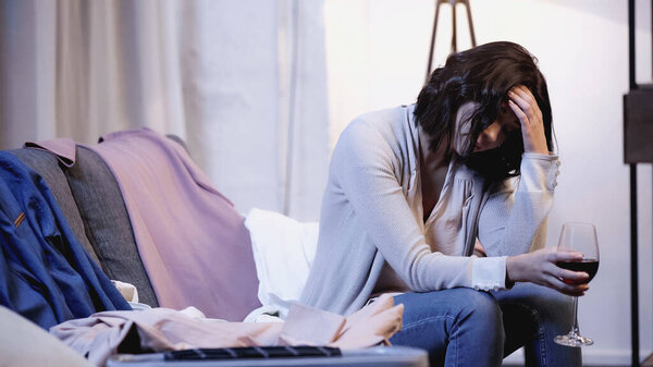 upset woman sitting on sofa with glass of red wine near male clothes and holding hand near head at home
