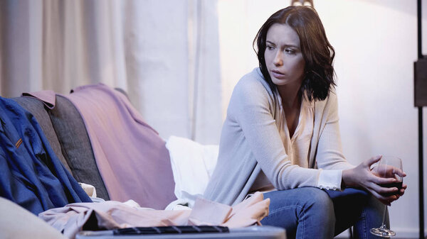 upset woman sitting on sofa with glass of red wine and looking at male clothes at home