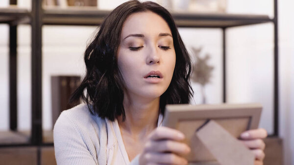 offended brunette woman looking at blurred photo frame in hands at home