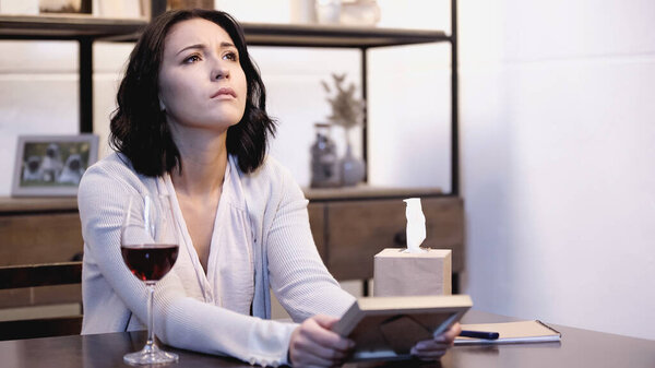 upset woman sitting on table with photo frame in hands and crying with tears at home