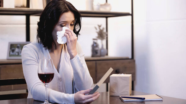 upset woman sitting on table, holding photo frame and wiping tears with paper napkin at home