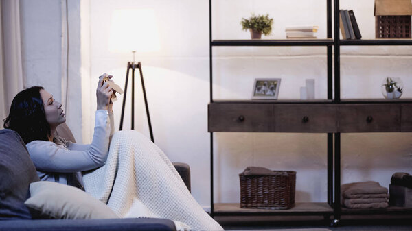 upset woman covered in blanket sitting on sofa and looking at photo frame in hands at home