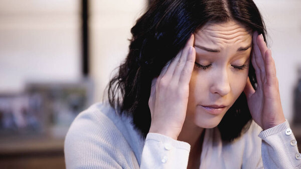 upset woman sitting with closed eyes and holding hands near head at home
