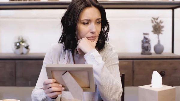 upset woman sitting on table with hand near face and holding photo frame at home