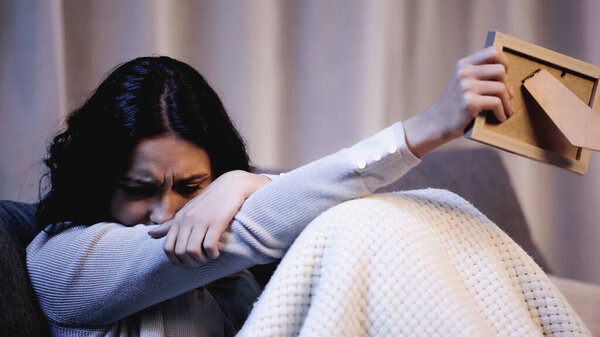 upset woman sitting on sofa and crying with outstretched hand holding photo frame at home