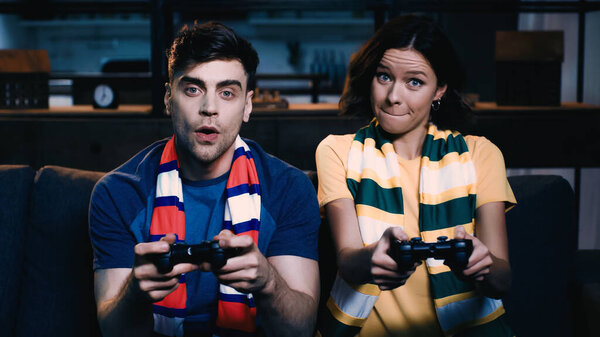 KYIV, UKRAINE - JUNE 09, 2021: worried couple in striped scarfs holding joysticks and playing video game 