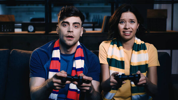 KYIV, UKRAINE - JUNE 09, 2021: stressed couple in striped scarfs holding joysticks and playing video game 