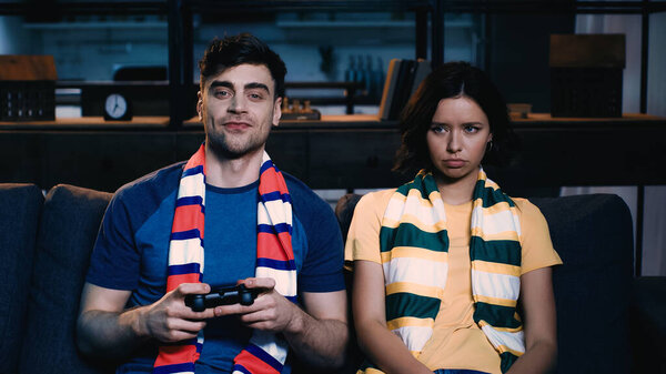 KYIV, UKRAINE - JUNE 09, 2021: cheerful man in striped scarf holding joystick and playing video game near offended girlfriend 