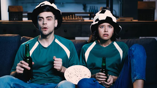 tensed man and woman in fan hats holding bottles of beer and watching championship in living room