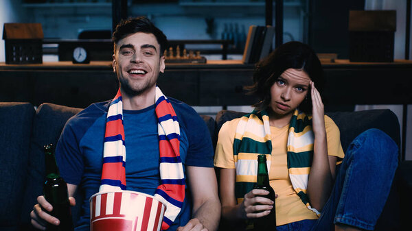 cheerful and upset sport fans holding bottles of beer near popcorn bucket while watching championship at home