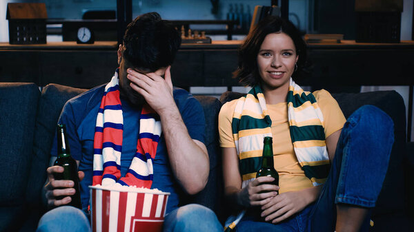 man covering face near woman smiling and watching sport match at home