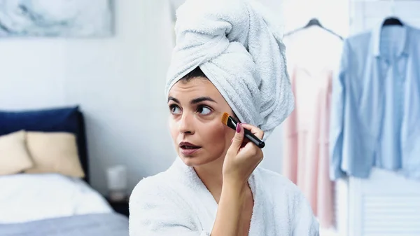 穿着浴衣 头裹毛巾的年轻女子在卧室用化妆品刷在脸上贴粉底 — 图库照片