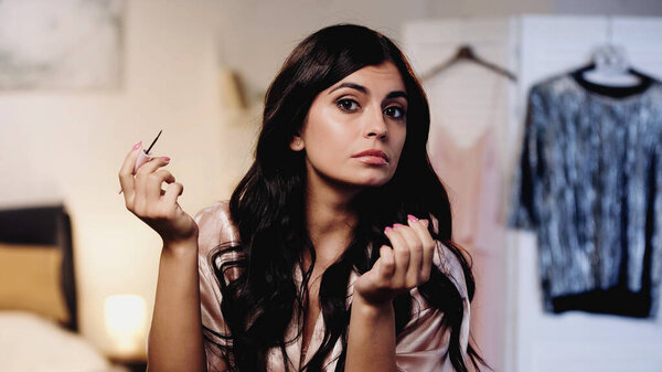 young woman in pink silk peignoir doing makeup with eyeliner in bedroom