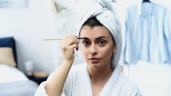 young woman in bathrobe with head wrapped in towel styling eyebrow with brush and looking at camera in bedroom