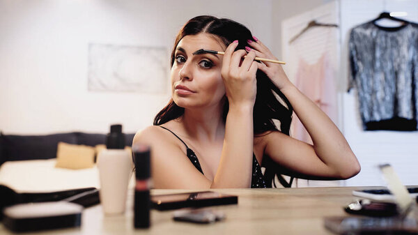 focused young woman adjusting hair and styling eyebrow with brush near table with decorative cosmetics in bedroom