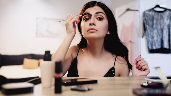 brunette young woman styling eyebrow with brush near table with decorative cosmetics in bedroom