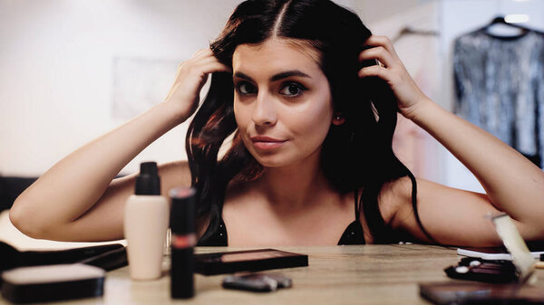 brunette young woman adjusting hair near table with decorative cosmetics in bedroom