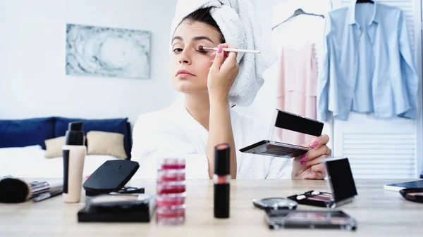 serious young woman with head wrapped in towel applying eye shadow with cosmetic brush in bedroom