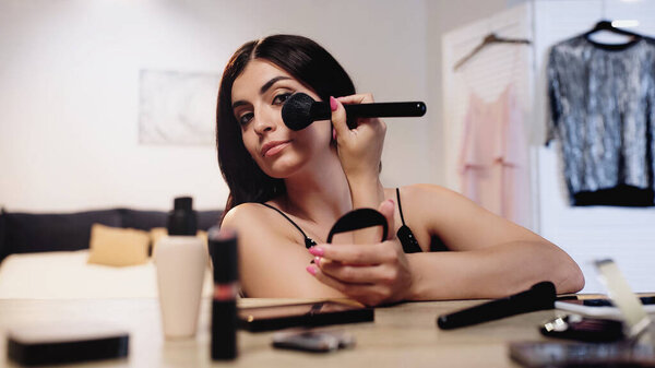 young woman applying face powder with cosmetic brush near table with decorative cosmetics in bedroom