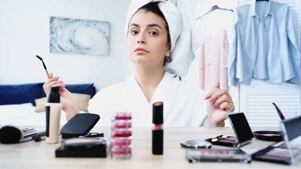 young woman sitting with eyebrow brush near table with decorative cosmetics in bedroom