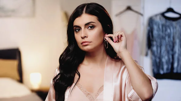 serious young woman in pink silk peignoir trying on earring in bedroom