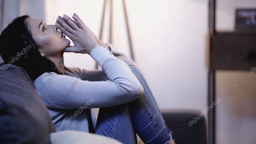 depressed woman in casual clothes sitting on sofa with praying hands near mouth and looking up at home