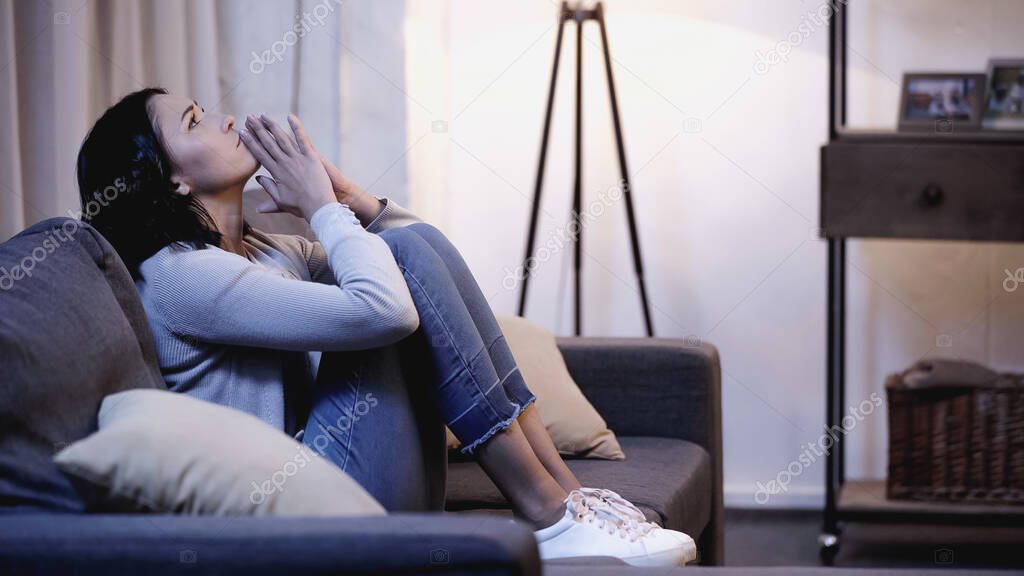 depressed woman in casual clothes sitting on sofa with praying hands near mouth at home